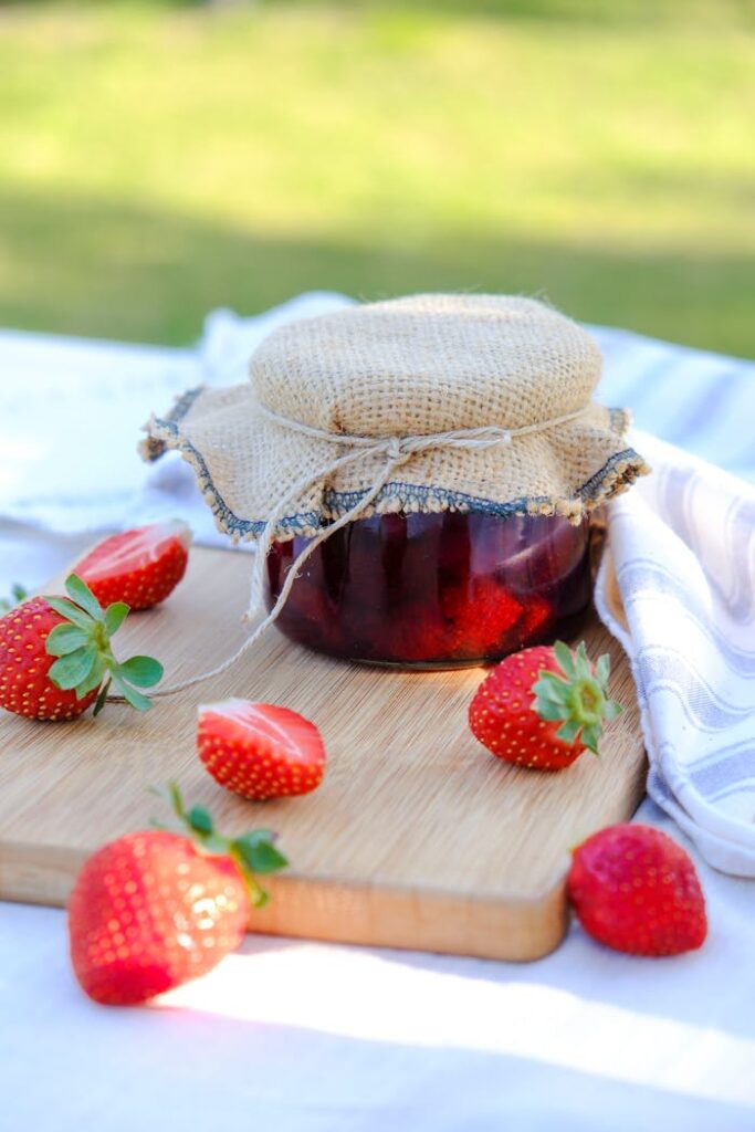 Jar of homemade strawberry jam with fresh strawberries on a wooden board at a picnic.