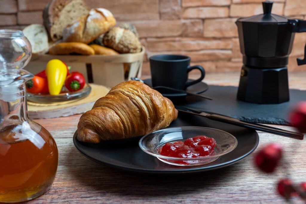A cozy breakfast setting featuring a croissant with jam, coffee, and a variety of bread.
