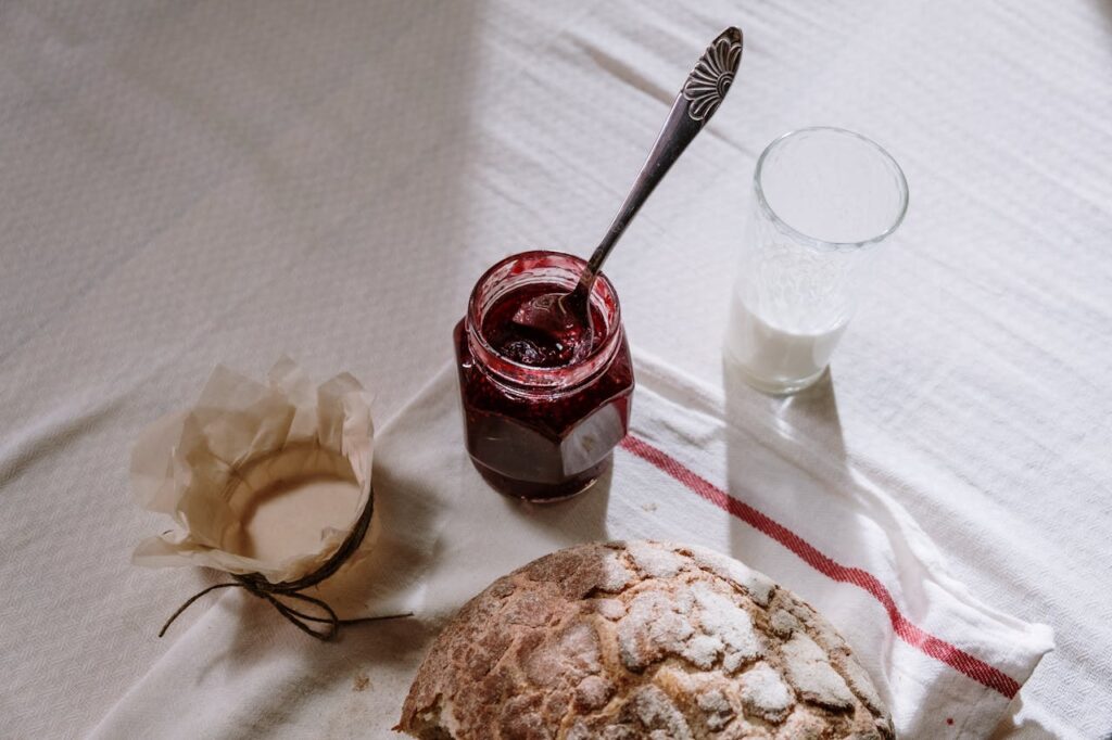 A cozy breakfast scene featuring rustic bread, a jar of jam, and a glass of milk on a textured tablecloth.