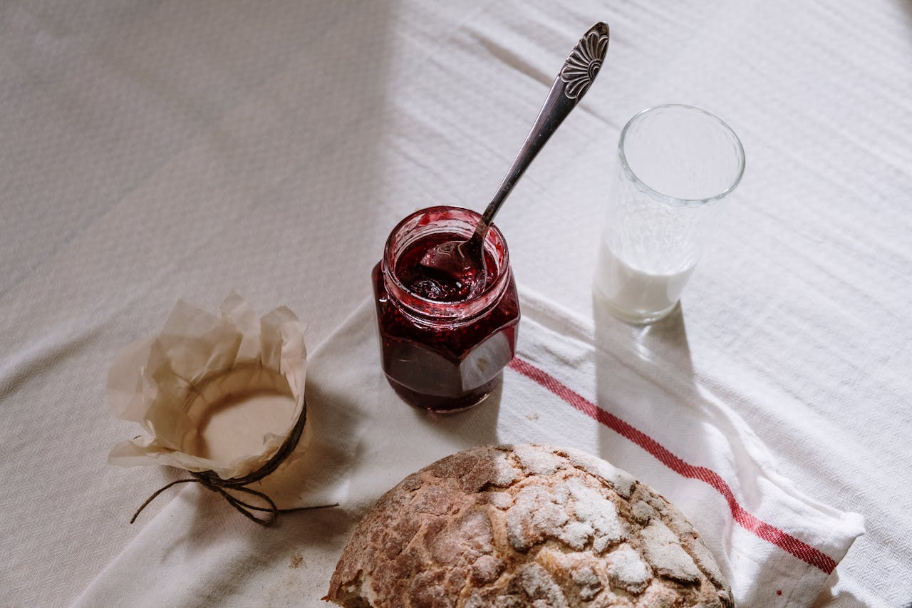 A cozy breakfast scene featuring rustic bread, a jar of jam, and a glass of milk on a textured tablecloth.